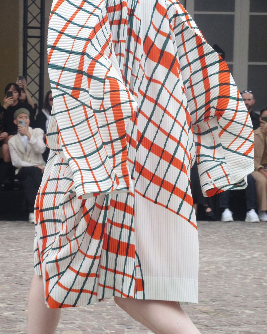 Fashion show attendee in a pleated, patterned dress featuring orange, green, and white, with other fashion show attendees blurred behind.