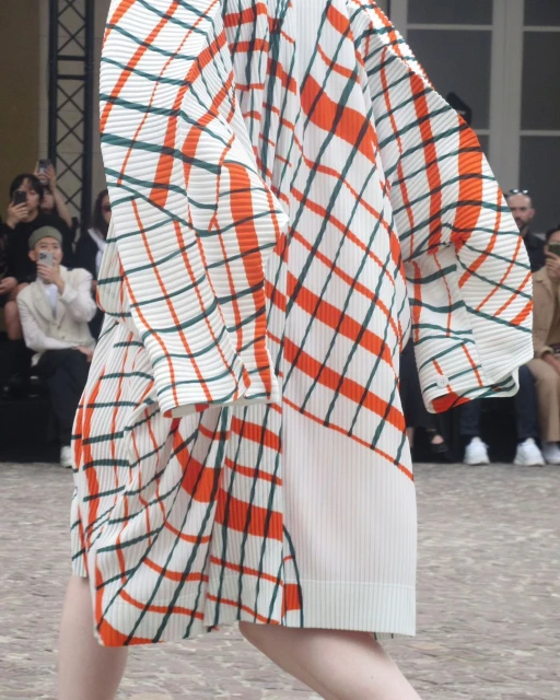 Fashion show attendee in a pleated, patterned dress featuring orange, green, and white, with other fashion show attendees blurred behind.
