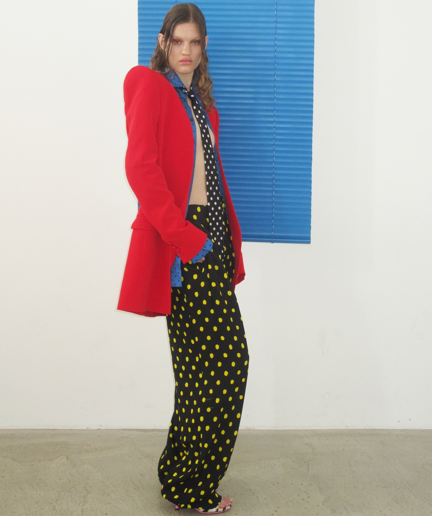 Fashionable woman in red blazer, patterned pants, and tie poses indoors against white wall with blue blinds, full shot, eye-level angle.
