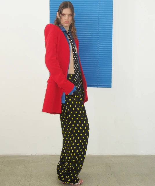 Fashionable woman in red blazer, patterned pants, and tie poses indoors against white wall with blue blinds, full shot, eye-level angle.