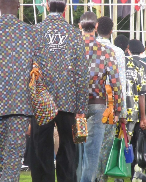 Fashion show featuring several men wearing checkered jackets with brand logos, accessorized with handbags in an outdoor setting. 
