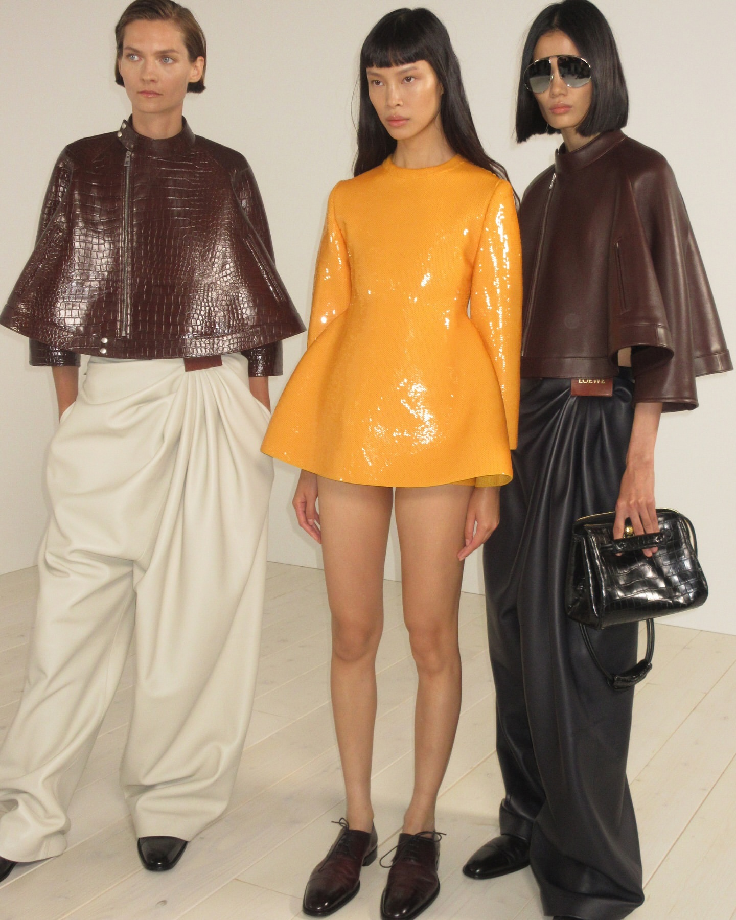 Three fashion models pose indoors, showcasing apparel; one in orange, two in brown, with beige and black trousers. Minimalist studio backdrop.