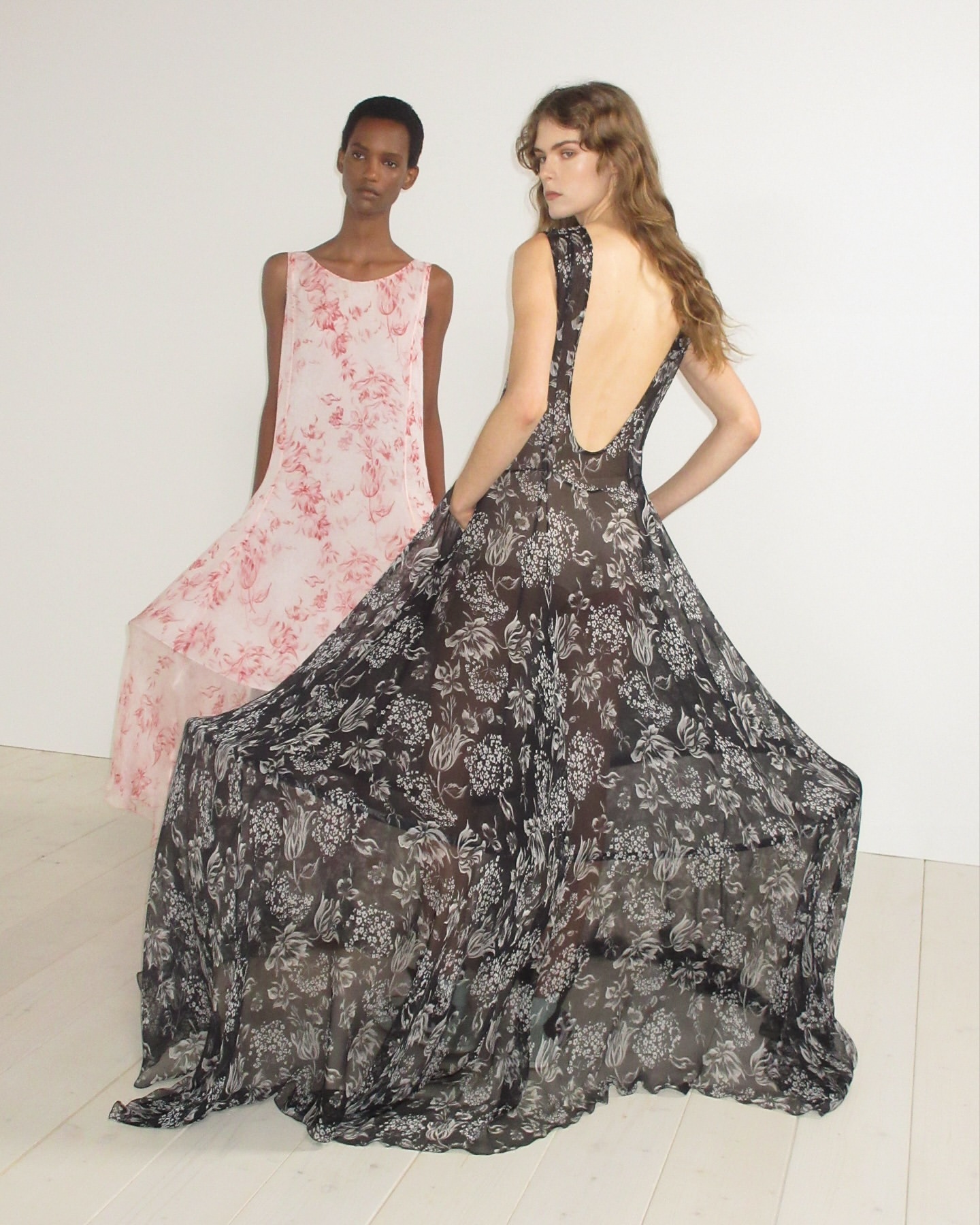 Two fashion models pose in flowing floral-print dresses against a white background in a studio setting, showcasing elegant and airy designs.