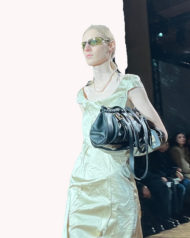 Runway model wearing a satin dress and carrying a black purse. The audience watches the fashion show from the background.