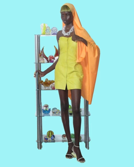 A dark-skinned female fashion model poses in a short yellow dress and orange headscarf next to a chrome shelving unit filled with glass art objects.