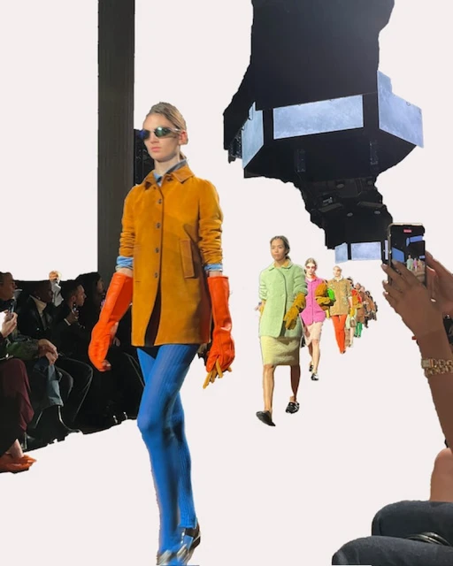 Runway fashion show featuring models in colorful outfits, captured mid-stride with an audience and photographers visible in the background.