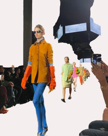 Runway fashion show featuring models in colorful outfits, captured mid-stride with an audience and photographers visible in the background.