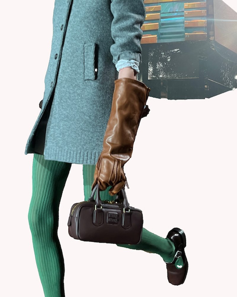 Fashion shoot featuring a model with green stockings, a gray coat, and a brown leather bag, posing against a neutral backdrop with an indistinct art piece.