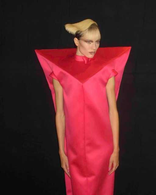 Woman in magenta gown with geometric collar and stylized blonde hair against a black background in a fashion portrait.