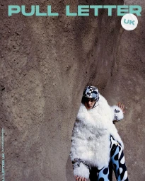 Model wearing a white faux fur coat and patterned trousers poses inside a rugged natural rock crevice.