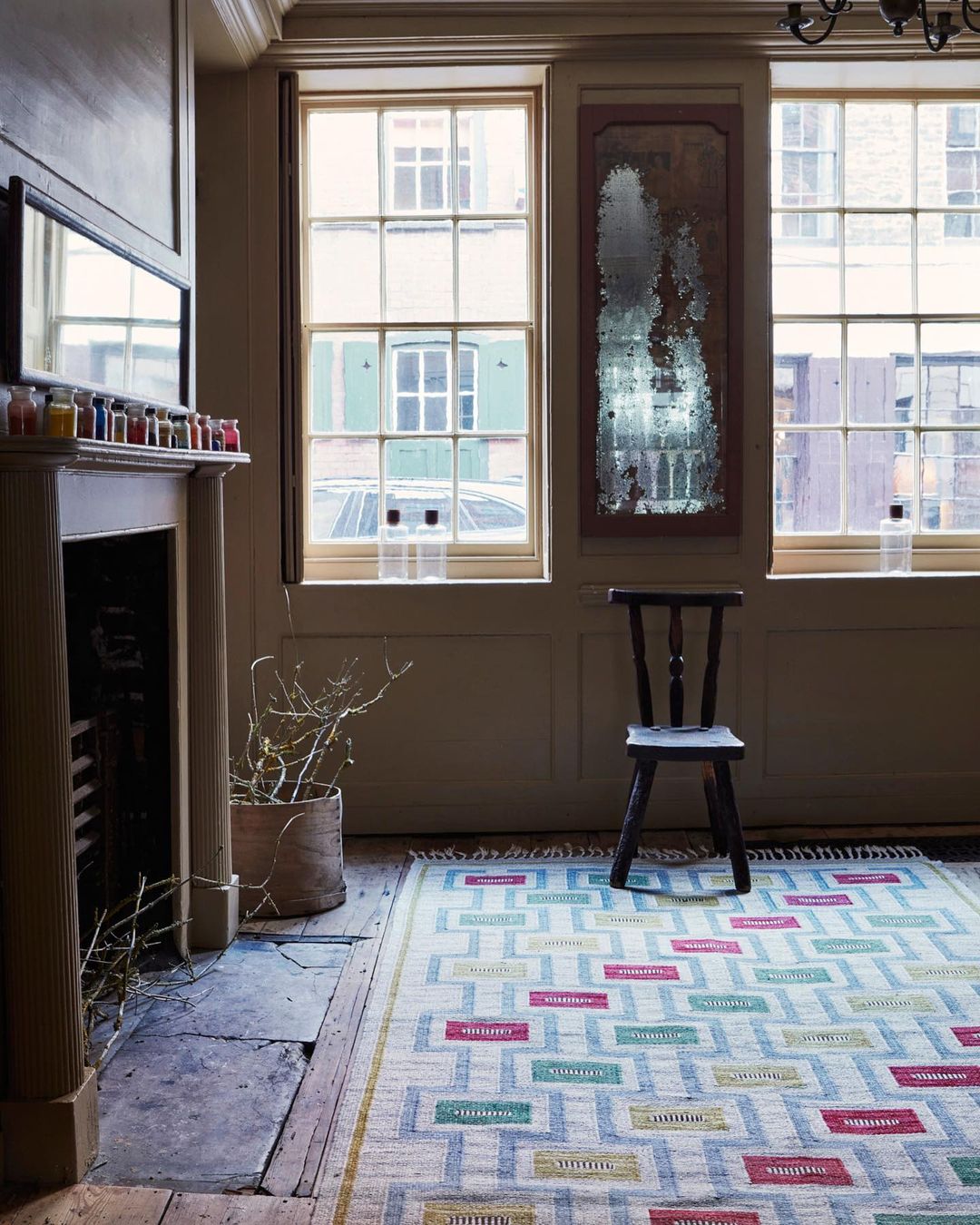 Interior view of a living room with a patterned rug, chair, fireplace, mantle with decorative jars, and natural light from windows.