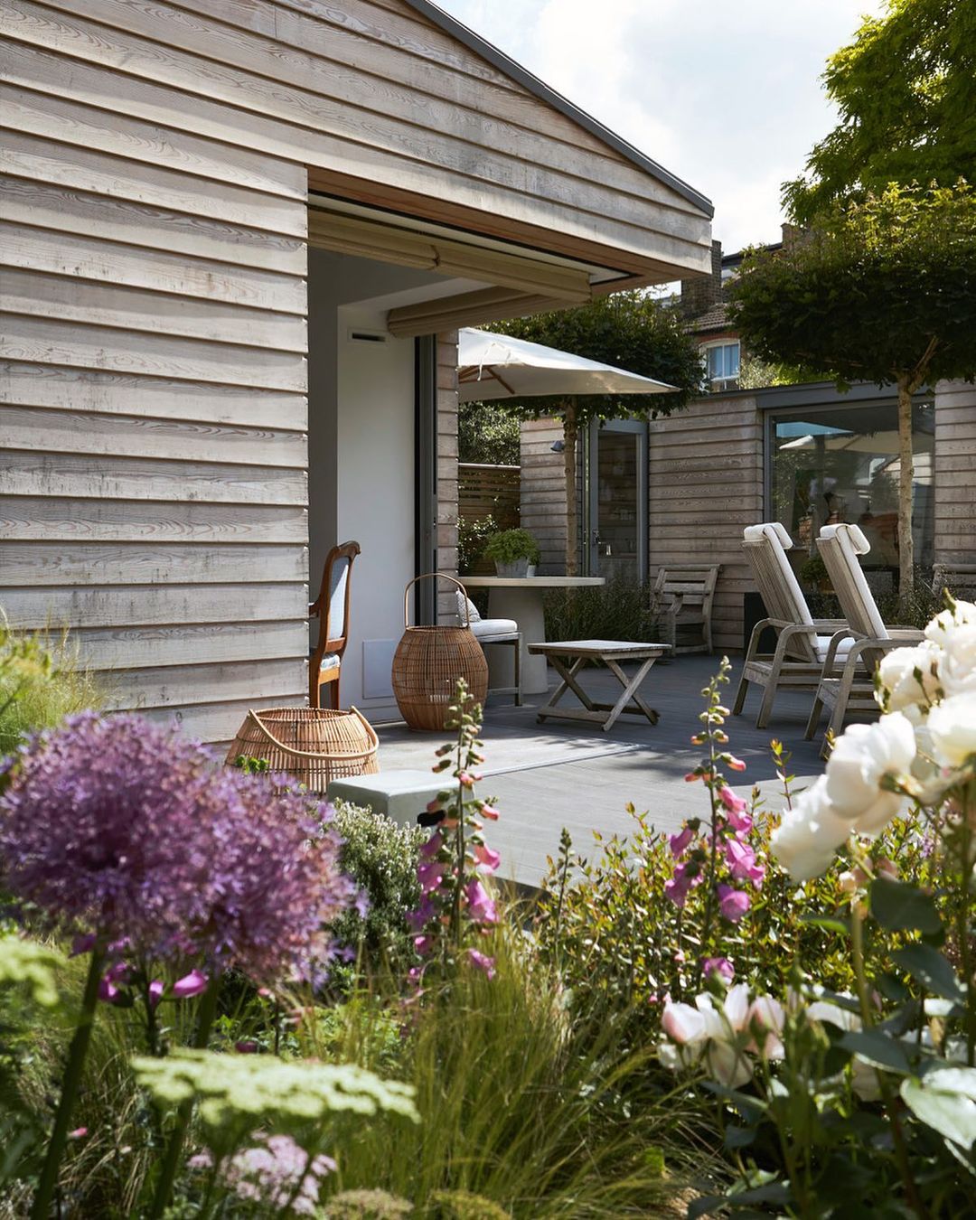 Exterior view of a modern house with wood siding, stone patio, outdoor seating, and flower garden featuring purple allium flowers.
