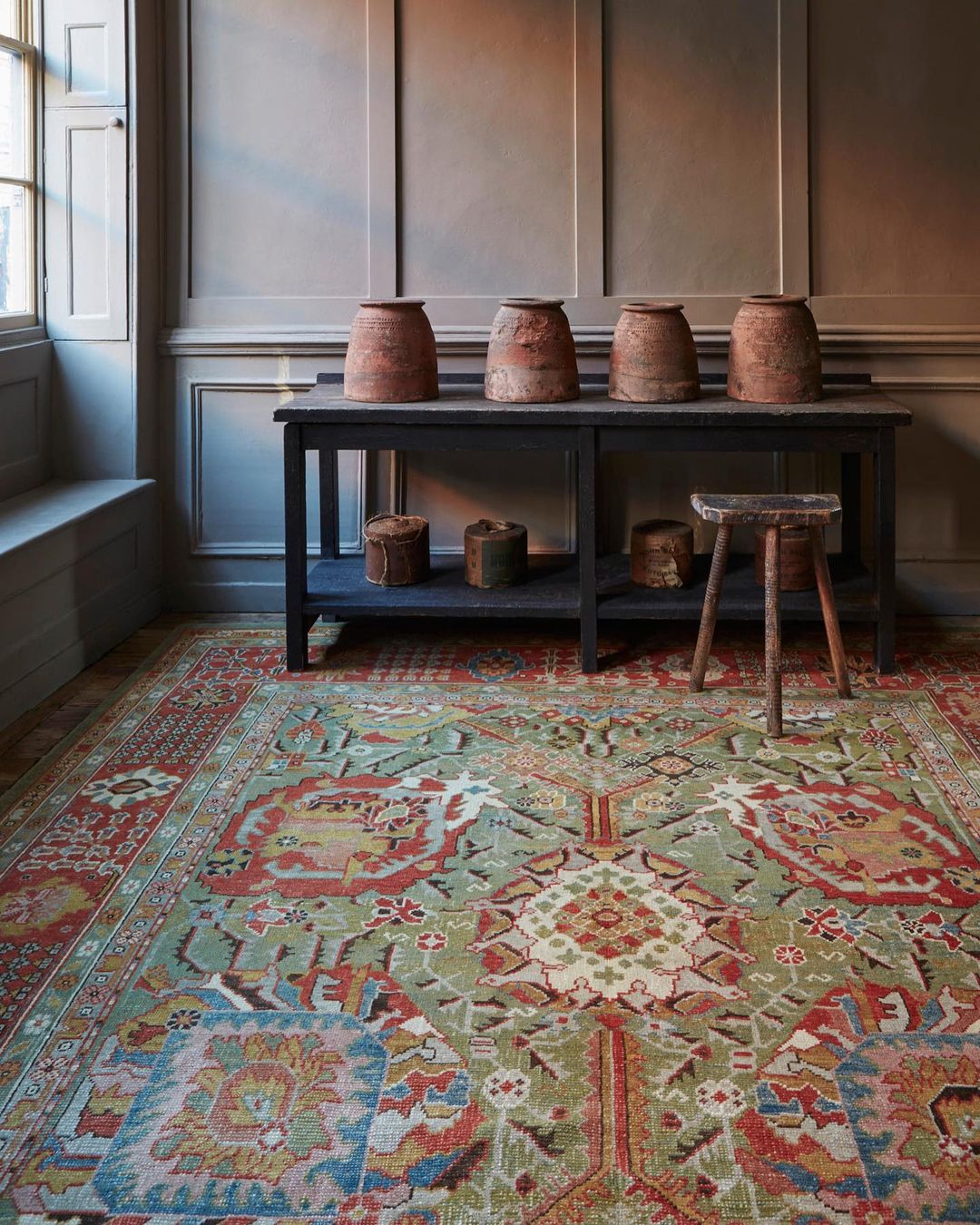 Interior of a room featuring a patterned rug, a black table with pots, and a stool creating a vintage and cozy home setting.