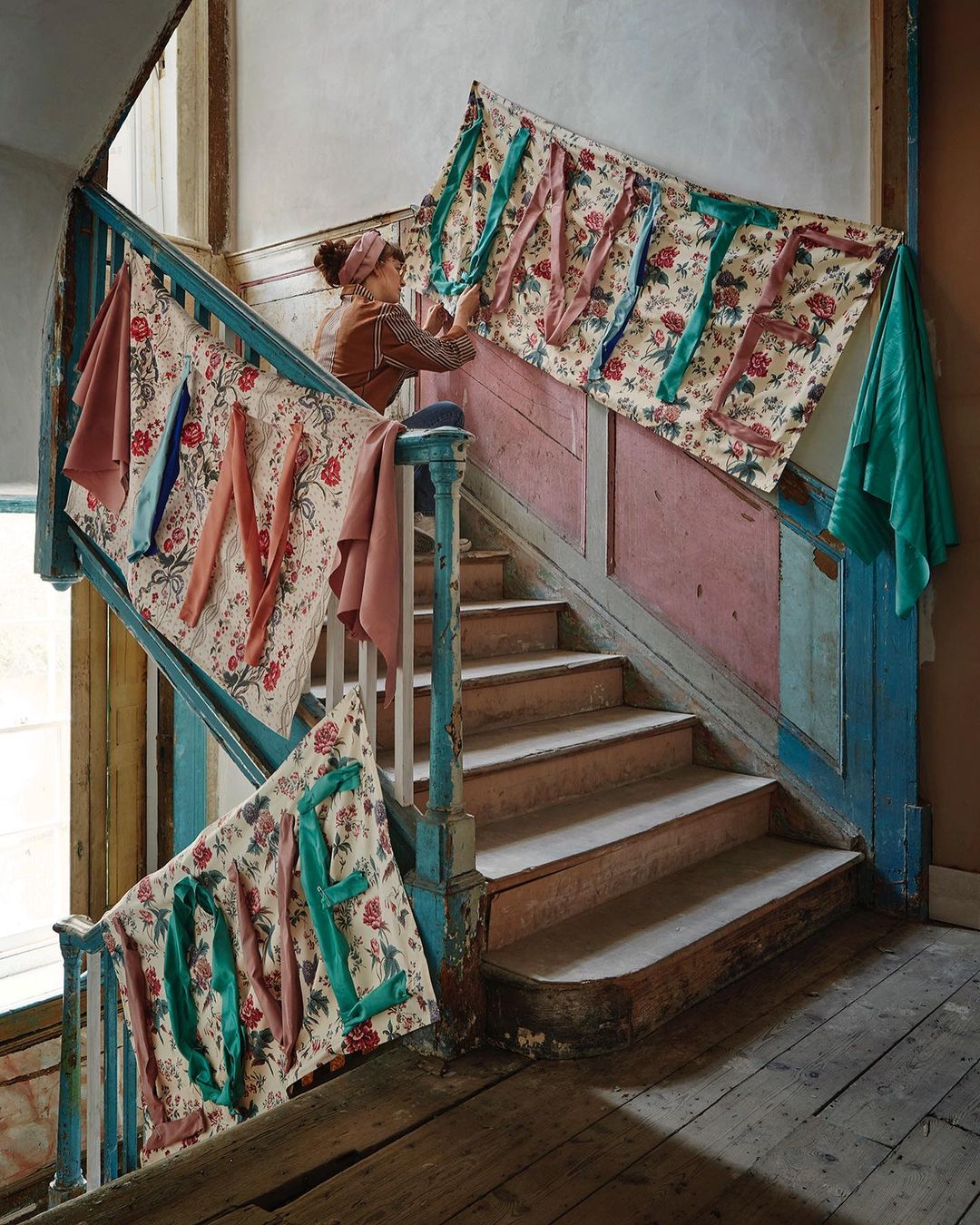 A woman stands on stairs decorated with floral fabric banners spelling 'WELCOME', creating a warm, inviting, and unique interior setting.