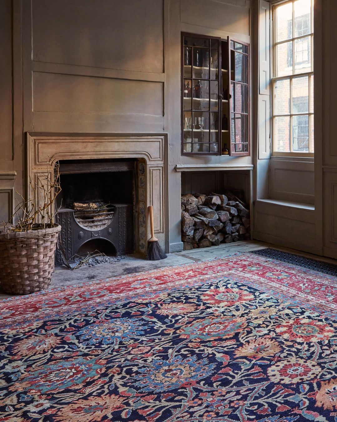 An ornate Persian rug fills an indoor space with a fireplace, wood storage, paneled walls, and natural light from a window.