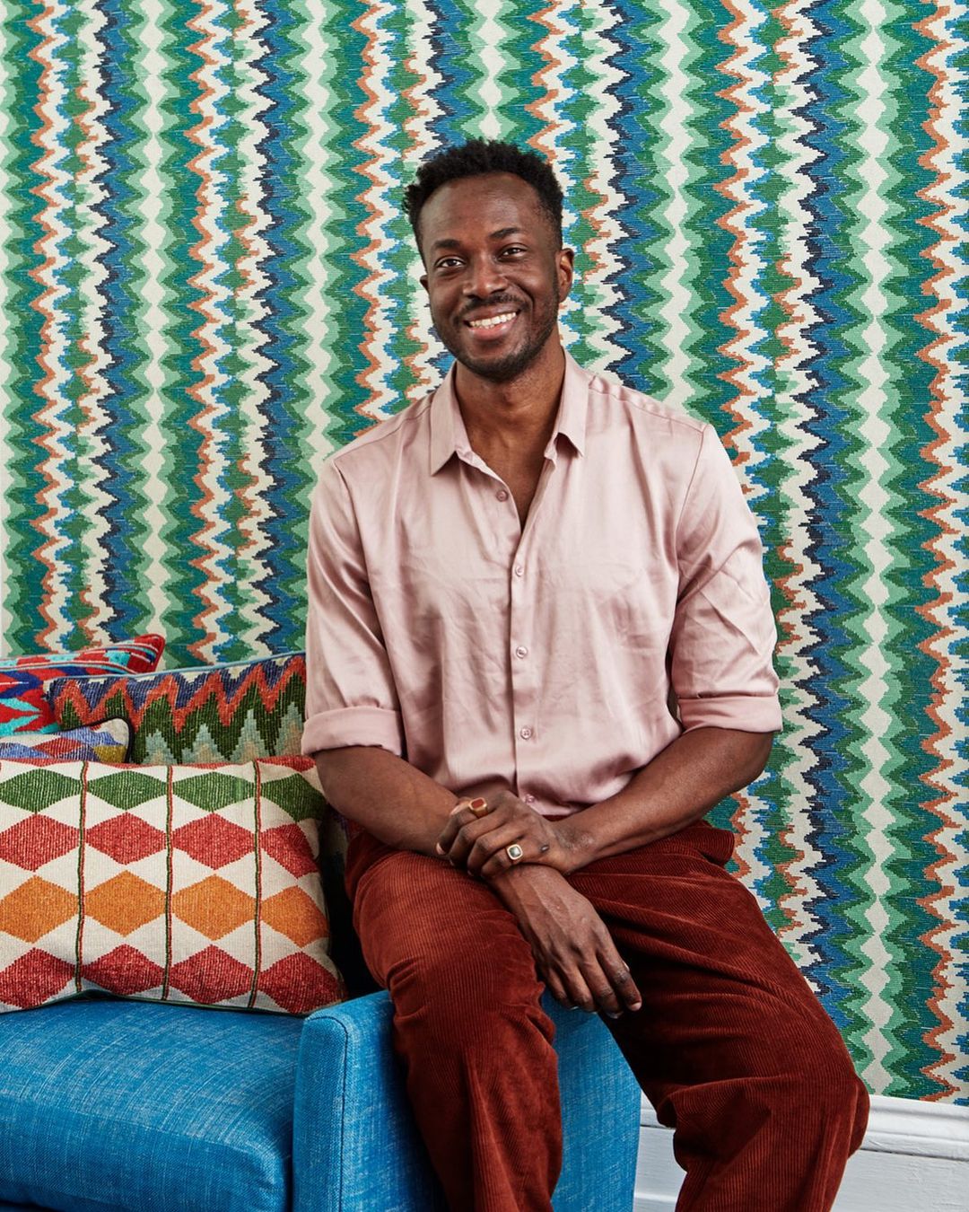 Smiling Black man sits on a blue chair in front of patterned wallpaper in a studio, wearing pink button-down shirt and reddish-brown pants.
