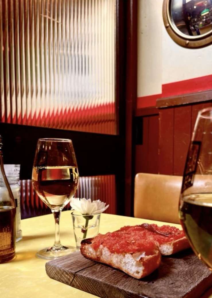 Still life features glasses of wine, bread with tomato, and a white flower in a glass on a yellow table with a textured glass background.