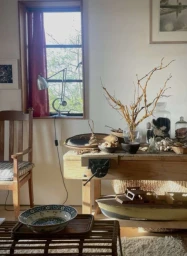 Rustic wooden workbench with dried branches in a glass vase, ceramic bowls, and a window with red curtains.