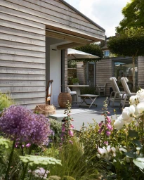 Modern wooden clad building exterior with patio, lounge chairs, and lush garden foreground by Jan Baldwin
