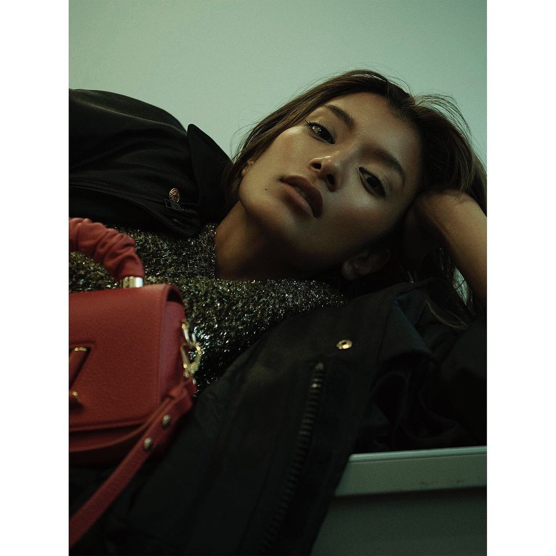 Portrait of a beautiful woman with dark hair wearing a metallic top, black jacket and holding a red purse in a low-angle shot.