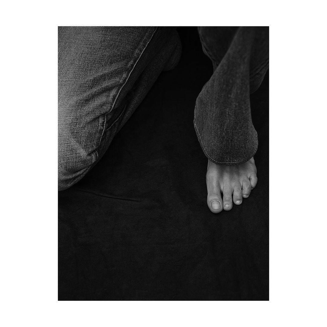 Black and white studio shot of person wearing jeans sitting with their foot extended forward on dark fabric background.