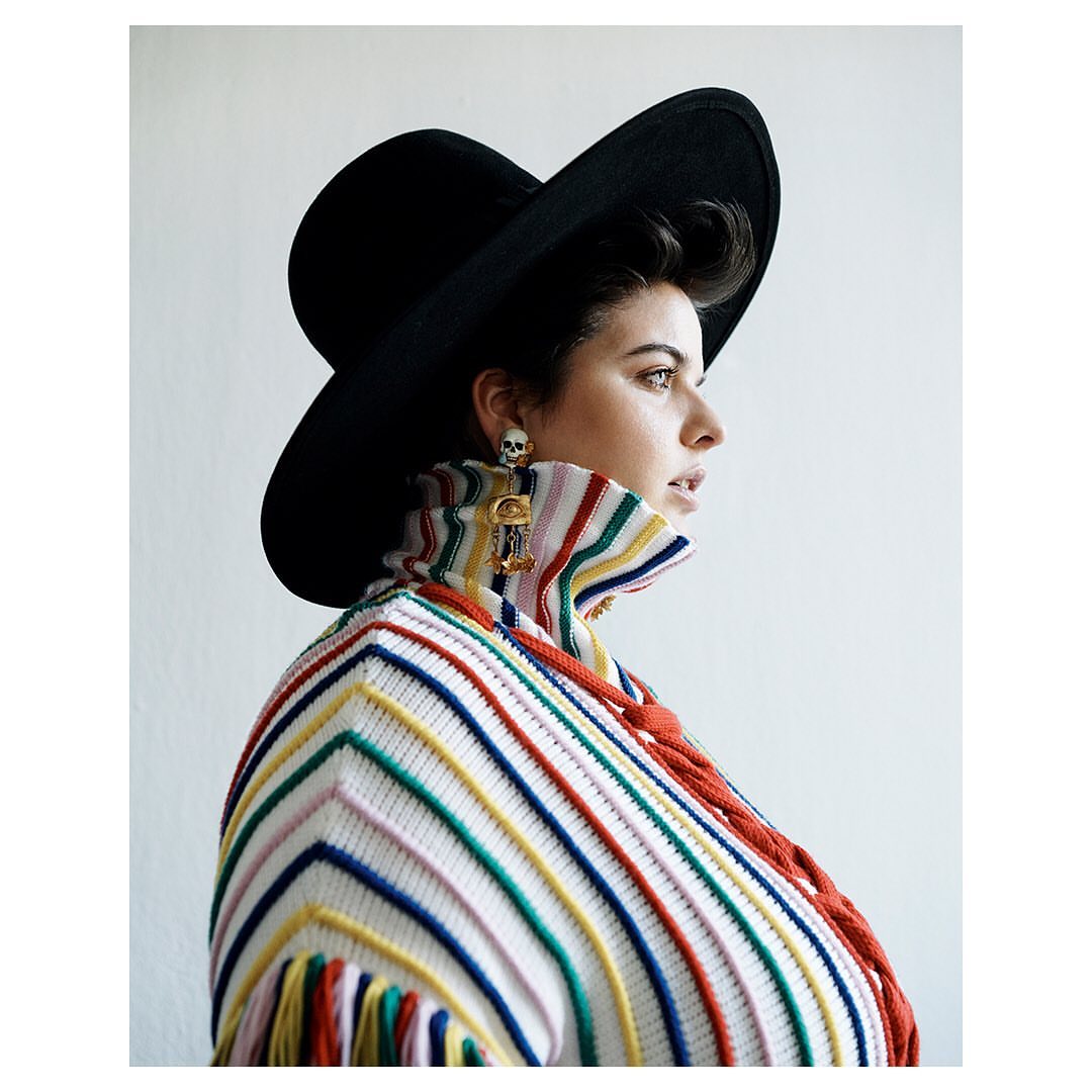 Portrait of a woman wearing a black hat and a striped sweater with fringe, posing against a white wall in a studio setting.