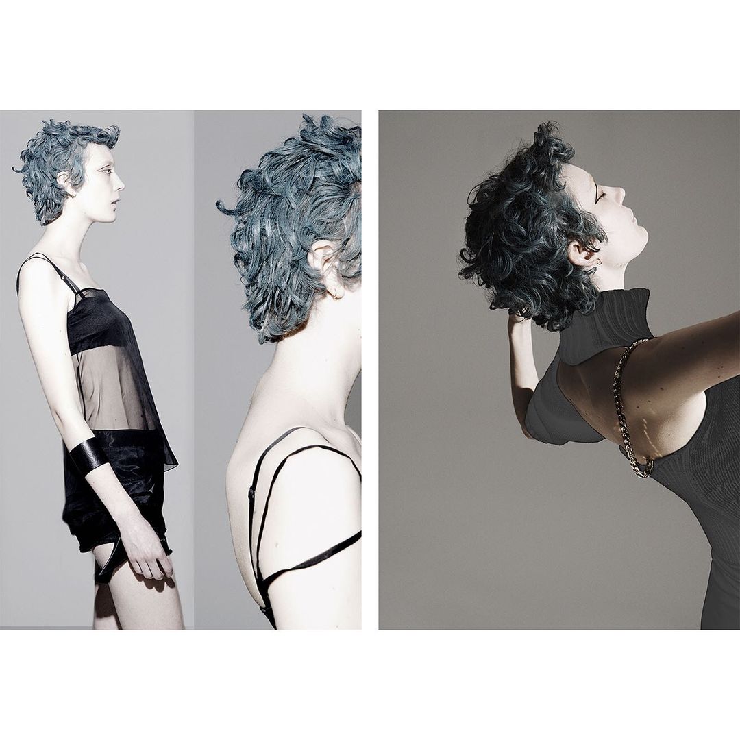 Fashionable young woman with blue hair in black outfit poses in a studio, showcasing a modern and edgy style against a grey background.