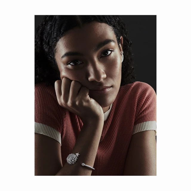 Close-up of a young woman with dark curly hair and a peach shirt, resting her face on her hand with two bracelets on her wrist.