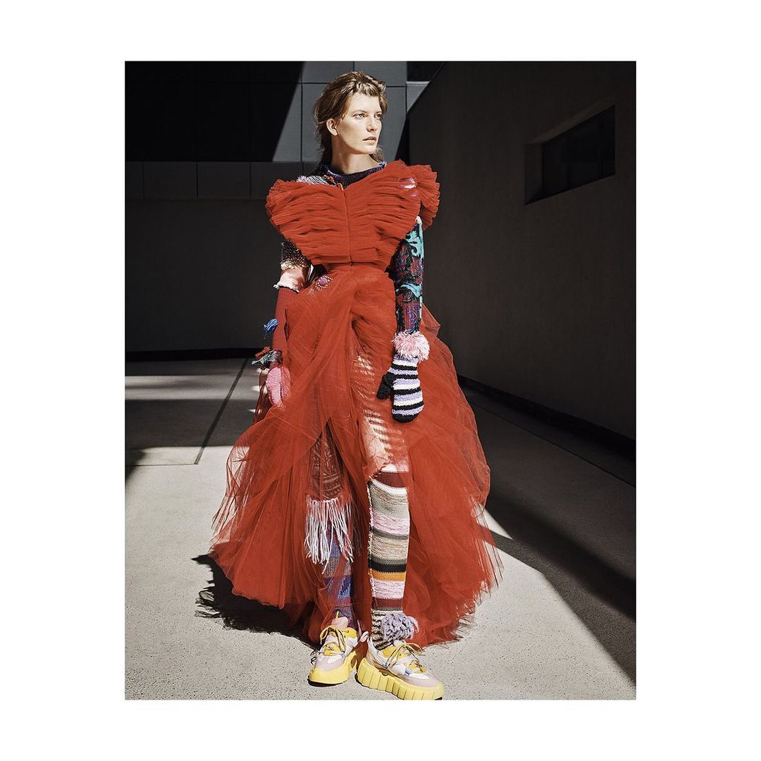 Editorial shot of a female model in a flowing red dress with textured leggings and sneakers posing against a concrete wall.