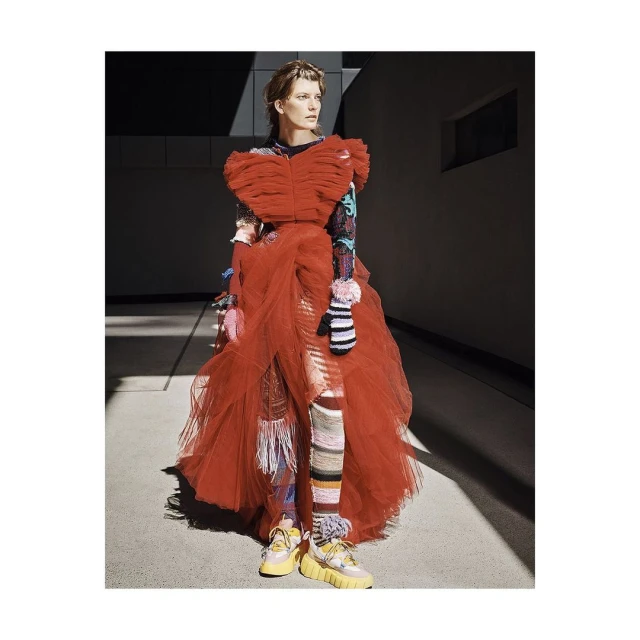 Editorial shot of a female model in a flowing red dress with textured leggings and sneakers posing against a concrete wall.