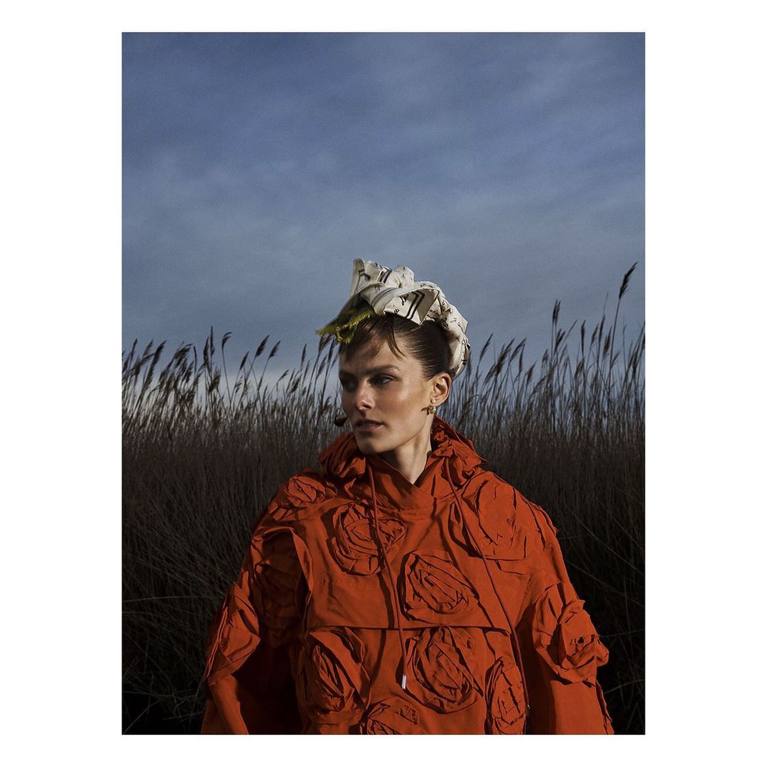 Woman in orange hooded jacket with floral hat stands amid tall grasses under a blue sky for a fashion or editorial shoot.