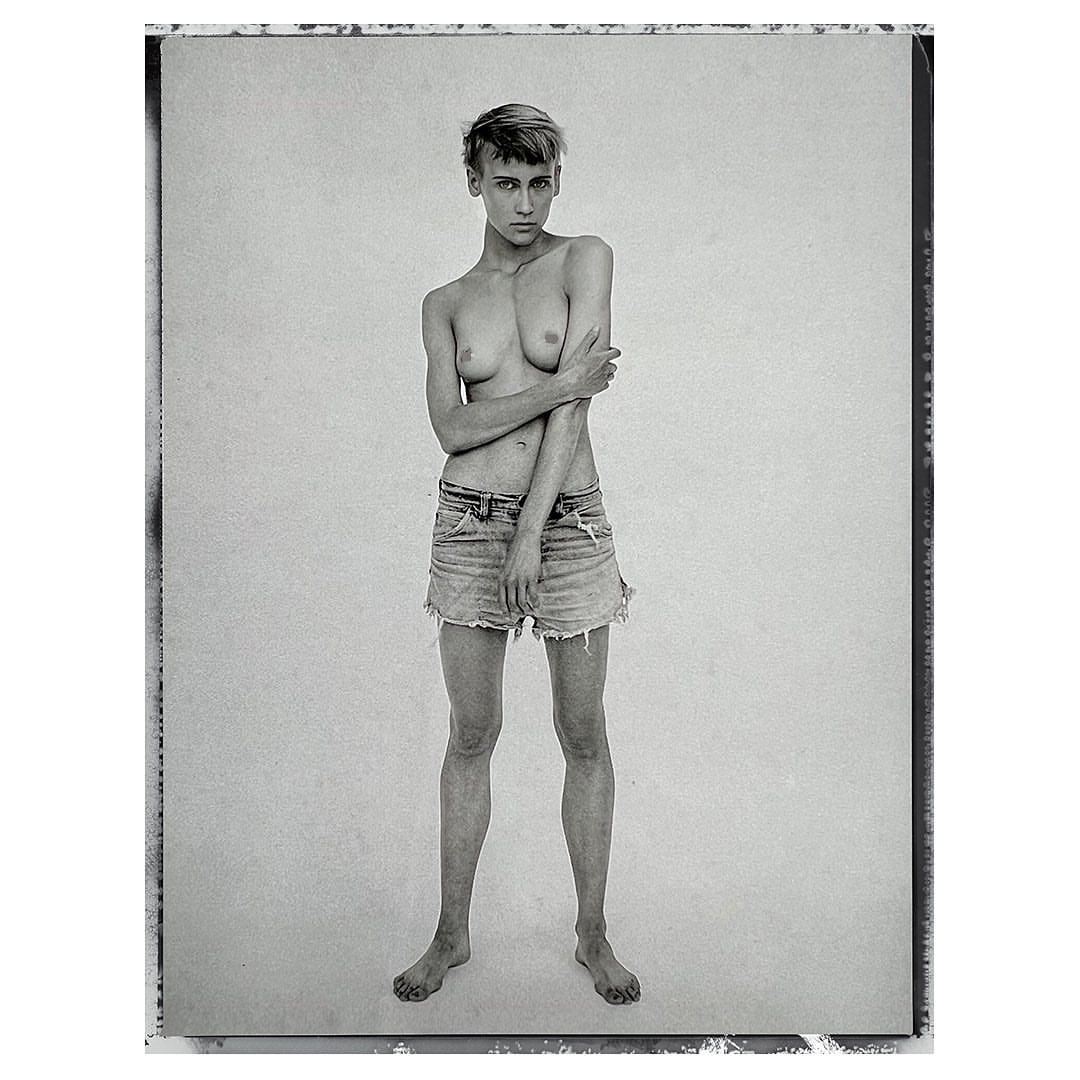 Black-and-white portrait of a topless young adult female in denim shorts, standing barefoot against a white backdrop in a photo studio.