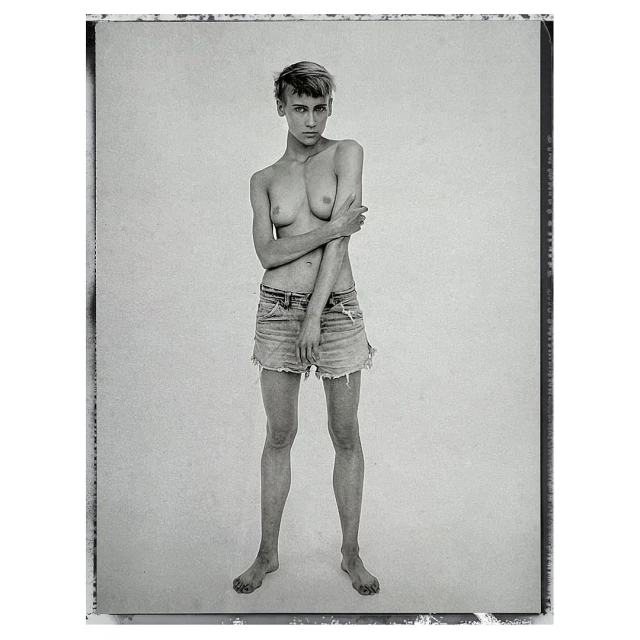 Black-and-white portrait of a topless young adult female in denim shorts, standing barefoot against a white backdrop in a photo studio.
