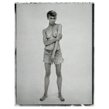 Black-and-white portrait of a topless young adult female in denim shorts, standing barefoot against a white backdrop in a photo studio.