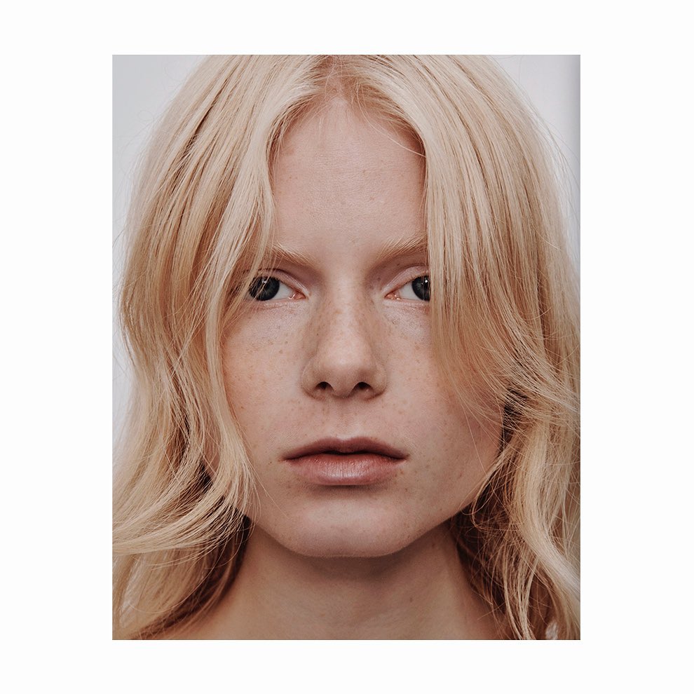 Close-up portrait of a young adult female with pale skin, freckles, and shoulder-length blonde wavy hair against a light grey background.