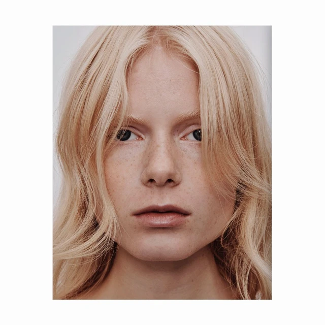 Close-up portrait of a young adult female with pale skin, freckles, and shoulder-length blonde wavy hair against a light grey background.