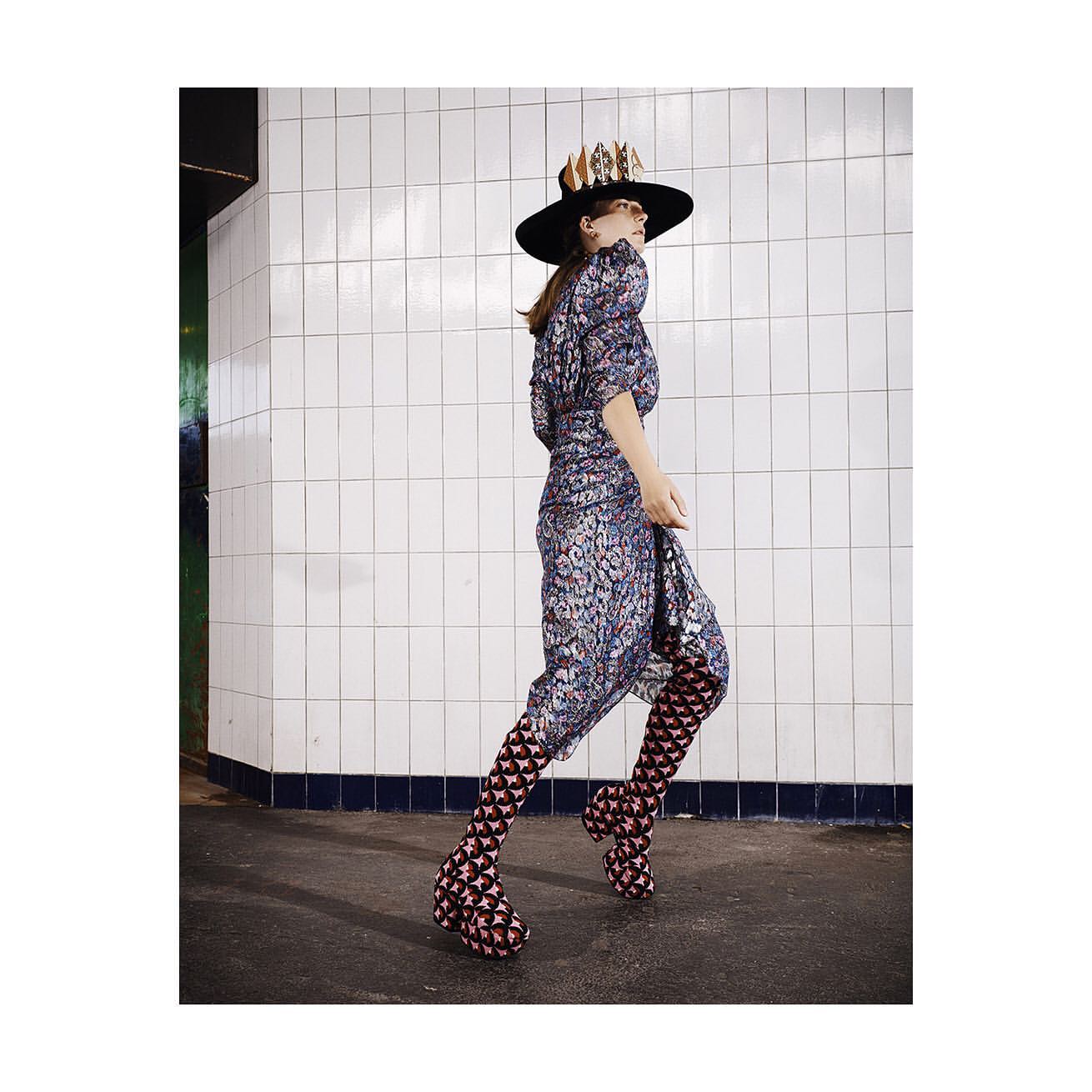 A woman in a patterned dress, black hat, and geometric patterned boots walks in front of white tiled wall in an underground setting.
