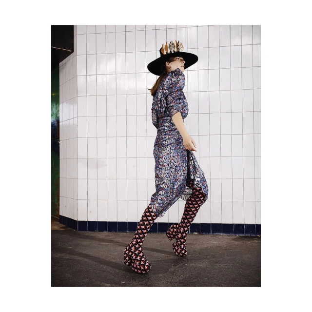 A woman in a patterned dress, black hat, and geometric patterned boots walks in front of white tiled wall in an underground setting.