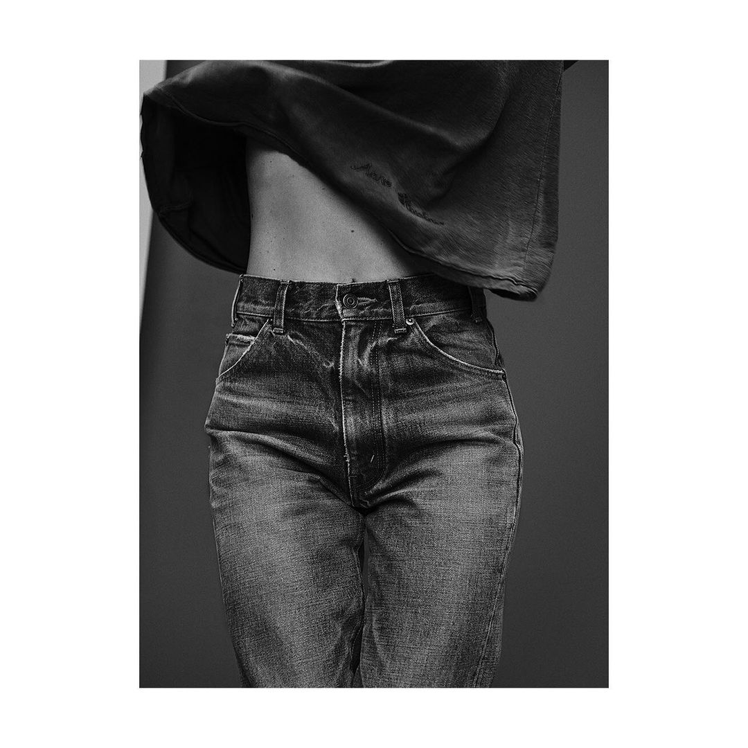 Black and white studio shot of adult woman's torso and jeans, low rise, with dark top partially concealing upper body and stomach.