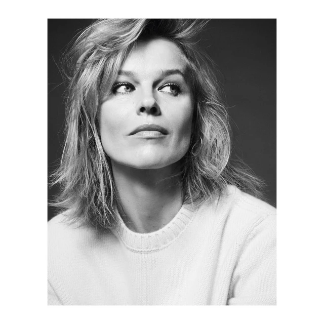 Black and white portrait of a blond woman wearing a white sweater looking upward with a subtle smile on a neutral background.