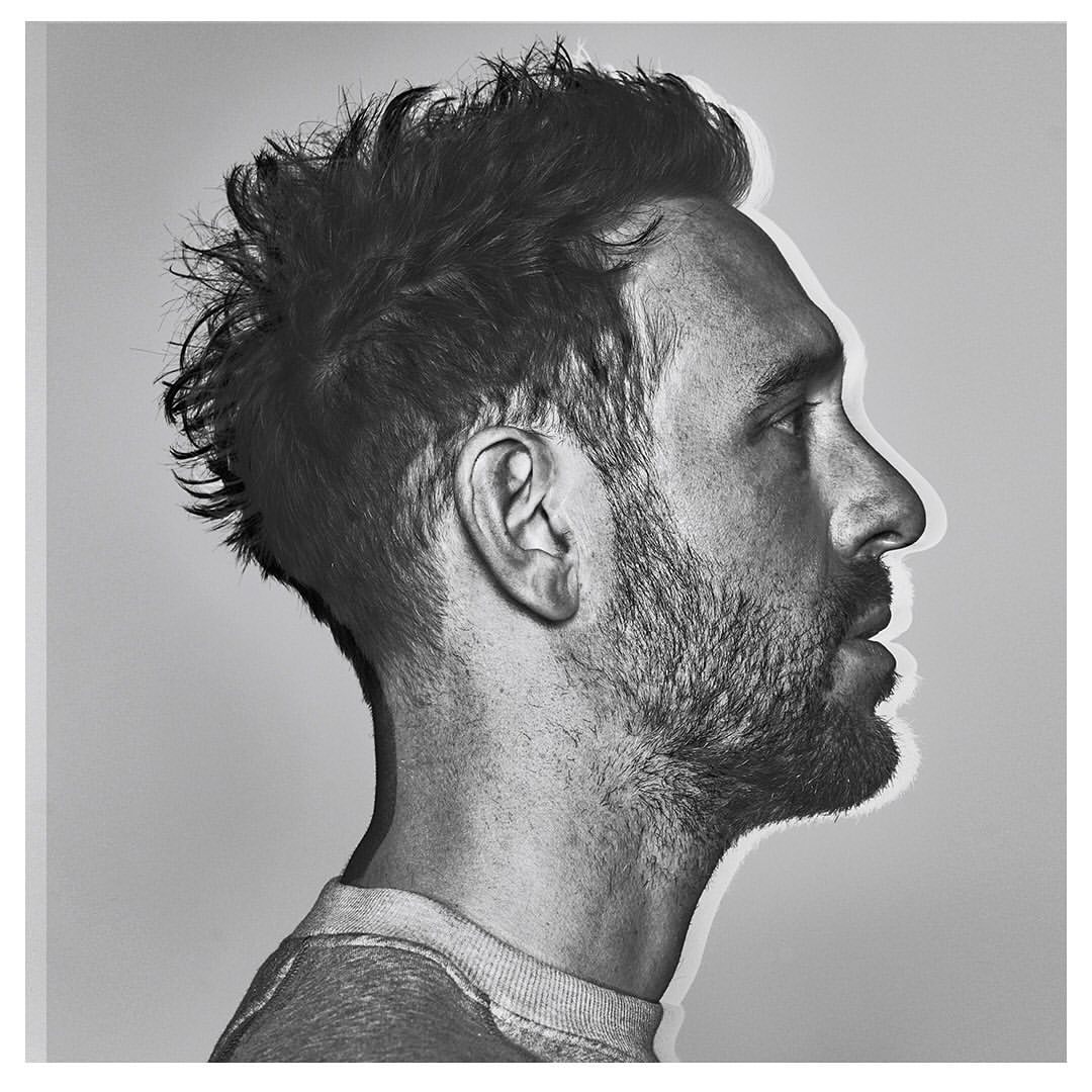 Monochromatic portrait of a light-skinned adult male in profile with messy dark hair, a beard, a grey sweatshirt, against a pale backdrop.