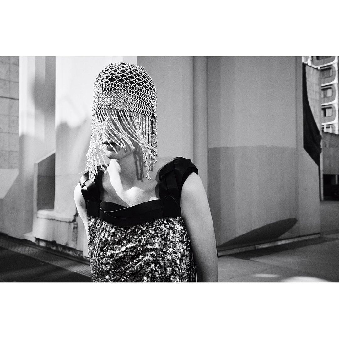 A woman poses outdoors in a black-and-silver dress and a silver beaded cap covering her face, with a neutral facial expression.