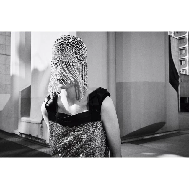 A woman poses outdoors in a black-and-silver dress and a silver beaded cap covering her face, with a neutral facial expression.