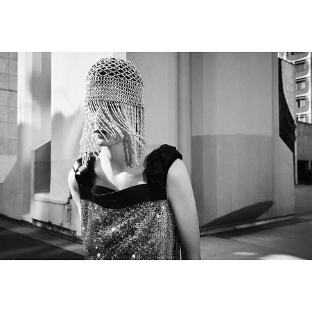 A woman poses outdoors in a black-and-silver dress and a silver beaded cap covering her face, with a neutral facial expression.
