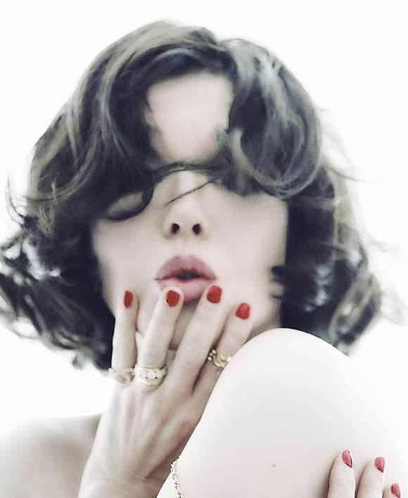 Woman with short wavy hair poses with red lipstick and red nail polish, wearing gold rings against a white backdrop in a beauty shot.