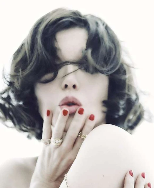 Woman with short wavy hair poses with red lipstick and red nail polish, wearing gold rings against a white backdrop in a beauty shot.