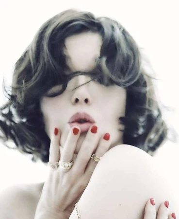 Woman with short wavy hair poses with red lipstick and red nail polish, wearing gold rings against a white backdrop in a beauty shot.