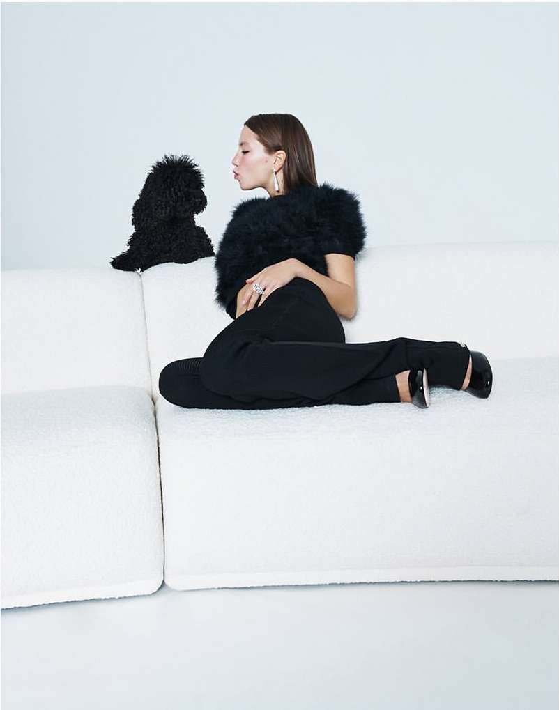Woman seated on a white couch, facing a black poodle. She wears black pants and top, with pale skin and brown hair.