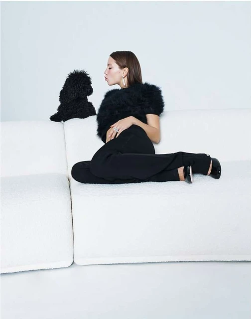 Woman seated on a white couch, facing a black poodle. She wears black pants and top, with pale skin and brown hair.