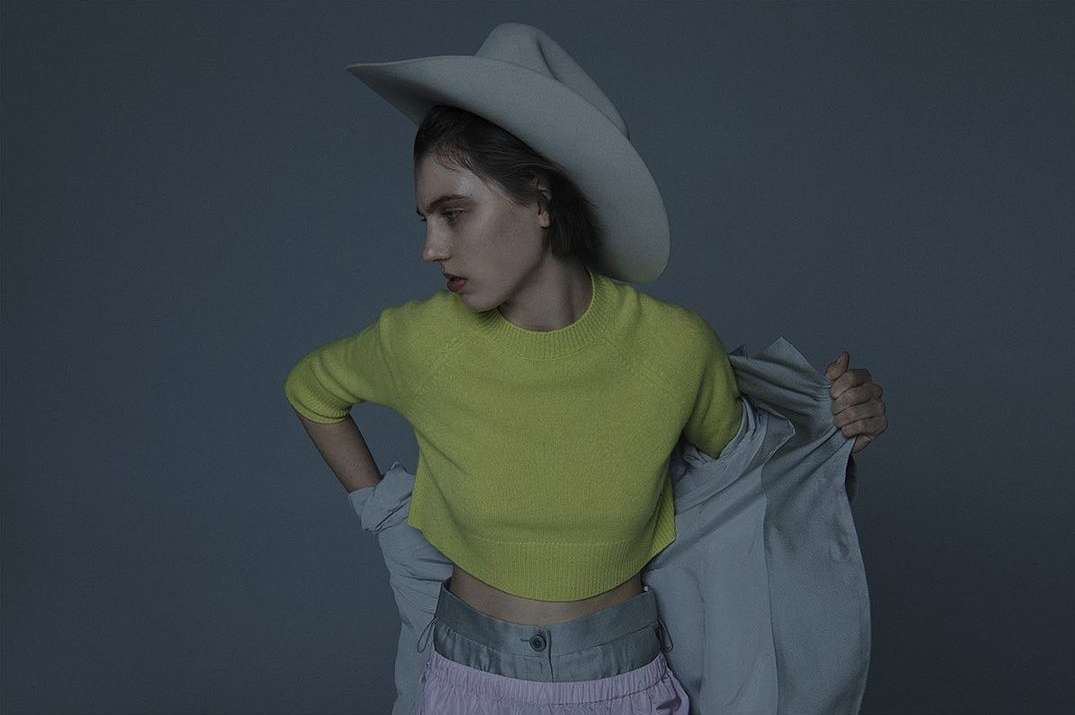 A fashion portrait of a young adult woman wearing a large hat, chartreuse top, and lilac pants in a photo studio setting.